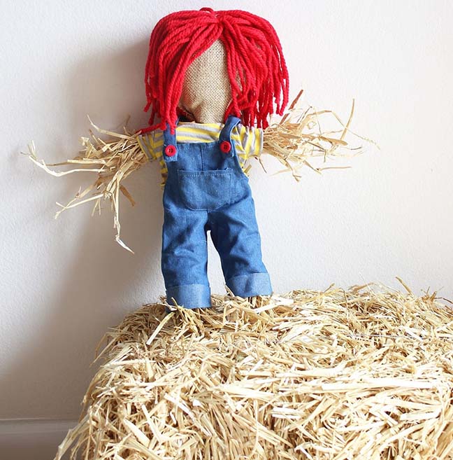 A doll dressed in overalls sits on top of hay.