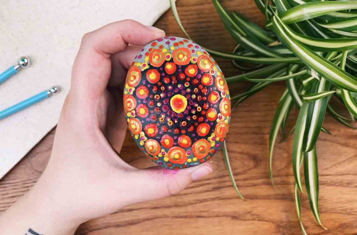 A hand holding a colorful egg on a table next to a plant.