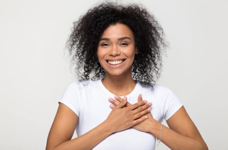 A young black woman with curly hair is holding her hands over her chest.