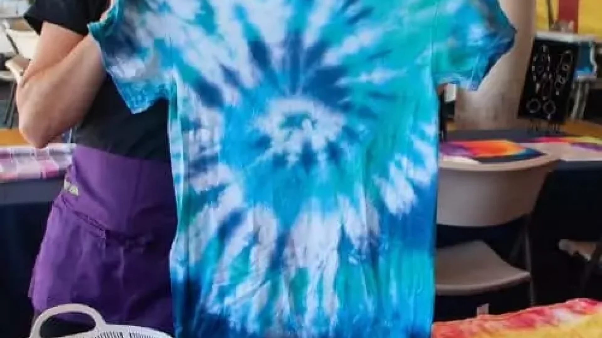 A woman holding a Kids Tie-Dye Party - San Diego Area t - shirt in front of a table.