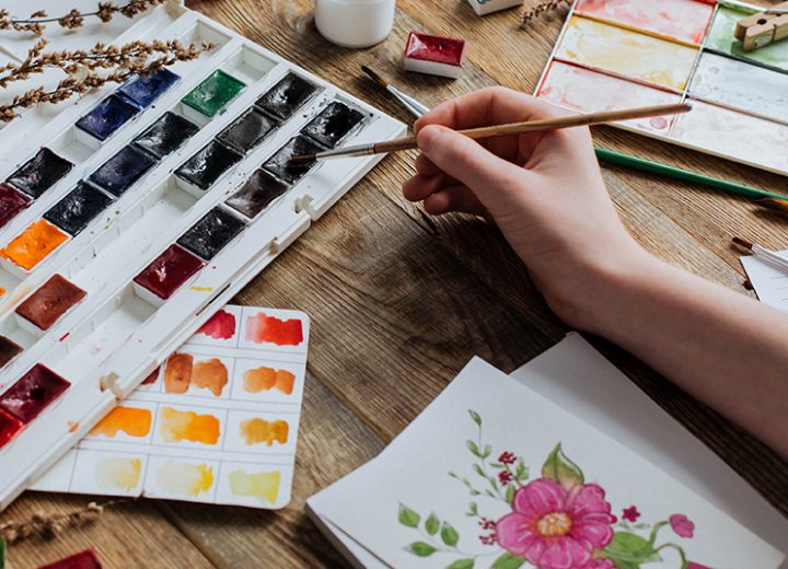 A young artist draws watercolors among paints and brushes on a wooden table.