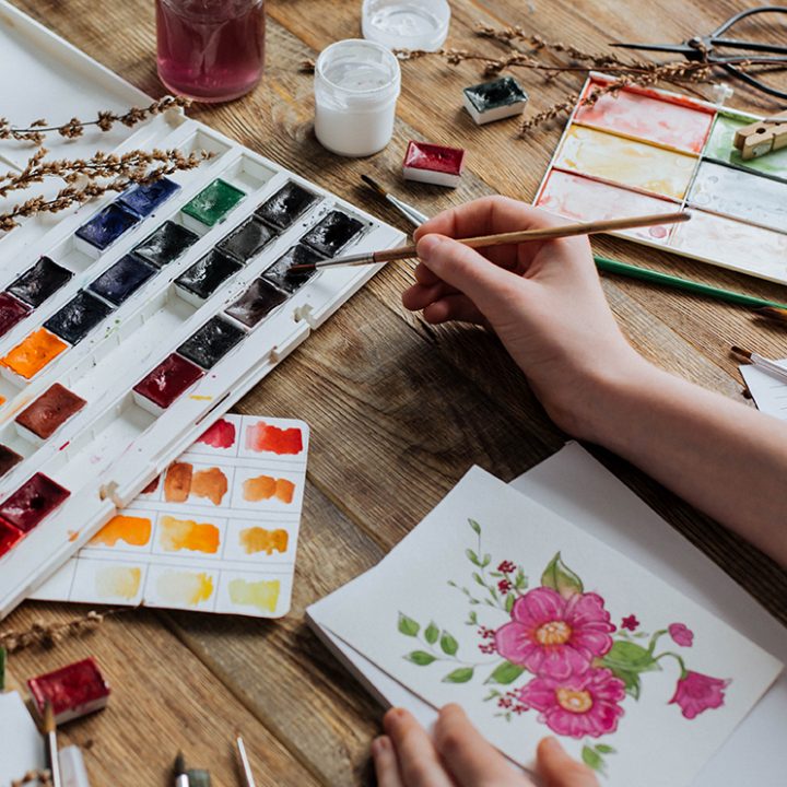A young artist draws watercolors among paints and brushes on a wooden table.